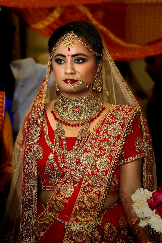 Beautiful Indian bride in a red and gold lehenga with intricate jewelry, posing elegantly indoors.